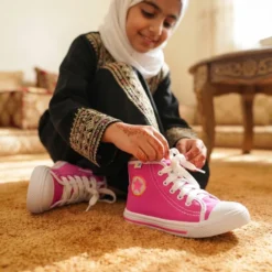 فتاة ترتدي حذاء كانفاس للفتيات 3F بينك ستار الفوشيا مع عباءة سوداء مطرزة وحجاب أبيض — تربط الأربطة على سجادة بيج